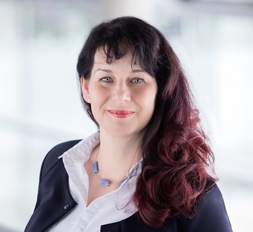 Sabine Vana-Ströhlas Kopf und Oberkörper sind zentral im Bild zu sehen. Sie trägt ein dunkles Sakko und ein helles Hemd. Ihre roten Haare fließen auf der rechten Seite um das Gesicht herum. Sie lächelt in die Kamera.